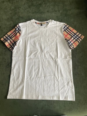 Vit t-shirt med rutiga ärmar Burberry - Snygg vit t-shirt från Burberry med klassiska beige, svarta och röda rutiga ärmar. T-shirten har rund hals och normal passform. Perfekt för dig som vill ha en stilren look med en twist. Tillverkad i mjuk bomull för skön känsla.