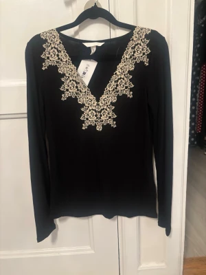 Viral tröja  - Snygg svart långärmad topp med v-ringning och dekorativa spetsdetaljer i beige runt halsen. Perfekt för dig som vill ha en stilren men ändå unik look. Toppen har en smickrande passform och känns både trendig och elegant.
