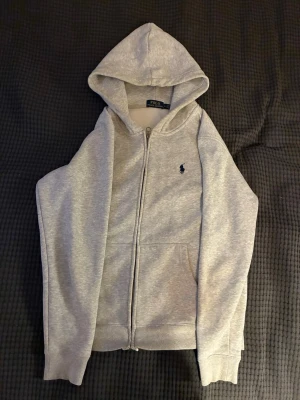 Grå zip hoodie från Polo Ralph Lauren - Snygg grå hoodie med dragkedja från Polo Ralph Lauren. Klassisk design med huva, fickor framtill och den ikoniska lilla loggan broderad på bröstet. Tillverkad i mjukt sweatshirtmaterial som är perfekt för chill dagar. Storlek M, har krympt lite i tvätten så skulle nog säga att den sitter mer som S.
