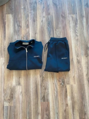  Les Deux - Säljer en mörkblå tracksuit från Les Deux med dragkedja på jackan och matchande byxor med dragsko i midjan. Båda delarna har Les Deux-loggan broderad på bröstet och benet. Tillverkad i mjukt material, perfekt för en avslappnad stil. 