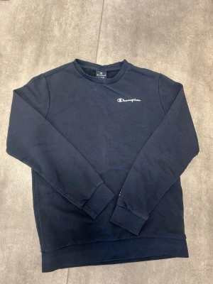 Mörkblå sweatshirt från Champion - Mörkblå sweatshirt från Champion med broderad logga på bröstet. Klassisk rund halsringning och ribbade muddar vid ärmslut och nederkant. Perfekt för en avslappnad och sportig stil.