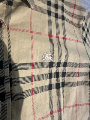Beige rutig skjorta med brodyr - Stilren beige skjorta med klassiskt rutmönster i svart och rött. Skjortan har lång ärm, knäppning framtill och en broderad logga på bröstet. Tillverkad i ett mjukt tyg som känns bekvämt mot huden. Perfekt för dig som gillar tidlös design.