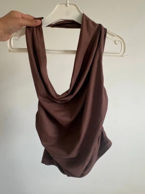 Brun draperad ärmlös topp - Stilren brun topp med draperad framsida och djup urringning. Toppen är ärmlös och har en lös passform som ger en avslappnad och trendig look. Perfekt att matcha med dina favoritjeans eller kjol.