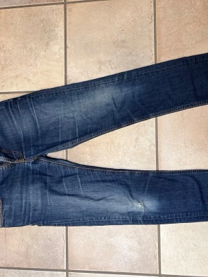 Mörkblå jeans från Nudie Jeans - Snygga mörkblå jeans från Nudie Jeans med klassisk femficksmodell och orangea kontrastsömmar. Jeansen har en smal passform och slitningar på låren och knät för en cool, använd look. Perfekta till vardagsstilen!