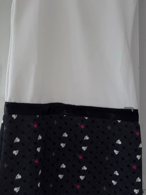 Vit och svart klänning med hjärtan - Snygg klänning med vit överdel och svart kjoldel dekorerad med små vita och rosa hjärtan. Kjoldelen har ett diskret rutmönster och ett svart band i midjan. Perfekt för dig som gillar söta detaljer och kontraster.str 152/158