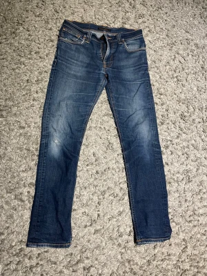 Blå raka jeans från Nudie Jeans - Säljer ett par klassiska blå jeans från Nudie Jeans med raka ben och snygga slitningar. Jeansen har fem fickor, kontrastsömmar och en cool detalj på bakfickorna. Perfekta till en avslappnad stil och tillverkade i denim. W32 L32