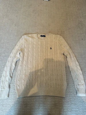 Beige kabelstickad tröja Polo Ralph Lauren - Kabelstickad tröja i beige från Polo Ralph Lauren med klassisk rund halsringning och broderad marinblå logga på bröstet. Stilren tröja utan problem som knappt är använd, hör av dig om frågor eller liknande OBS! Står S men passar även mot M hållet