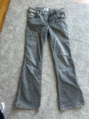 Grå bootcut jeans med fickdetaljer - Säljer ett par grå bootcut jeans med snygga ficklock och knappar bak. Jeansen har klassisk femficksmodell och är i ett mjukt denimtyg. Perfekta för dig som gillar en avslappnad men ändå trendig look.