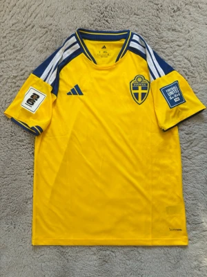 Sverige hemmatröja 2026 - Gul svensk landslagströja från Adidas med blå detaljer och vita ränder på axlarna. Tröjan har broderad svensk landslagssköld på bröstet och tryck på båda ärmarna. Tillverkad i funktionsmaterial (Climacool) för sport.