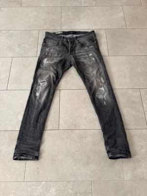 Grå slitna jeans från Replay - Säljer ett par grå jeans från Replay med slitningar och hål på benen för en cool, avslappnad look. Jeansen har fem fickor, smal passform och detaljerade sömmar. Perfekta för dig som gillar streetstyle och vill ha ett par trendiga jeans.