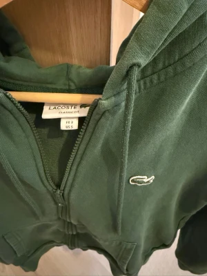 Grön zip hoodie från Lacoste - Snygg grön hoodie från Lacoste med dragkedja framtill och klassisk krokodil-logga på bröstet. Modellen har huva med dragsko och stora fickor framtill. Tillverkad i mjuk bomullsblandning och har ribbade muddar vid ärmar och nederkant.