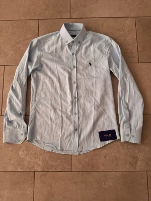 Ljusblå skjorta från Polo Ralph Lauren - Klassisk ljusblå skjorta från Polo Ralph Lauren med broderad logga på bröstet. Skjortan har lång ärm, knappar hela vägen och button-down krage. Perfekt för en stilren look.
