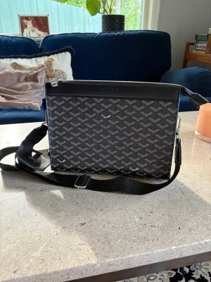 Svart axelväska med mönster - Snygg svart axelväska med justerbar rem och silverfärgad dragkedja. Väskan har ett grafiskt mönster i grått och insidan är fodrad i gult tyg. Perfekt storlek för det viktigaste och har en innerficka för småsaker.