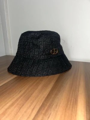 Svart bucket hat med monogram - Snygg svart bucket hat med diskret monogrammönster och guldfärgad detalj på sidan. Hatten har en klassisk form och är tillverkad i ett tyg med struktur. Perfekt accessoar för att lyfta din outfit.