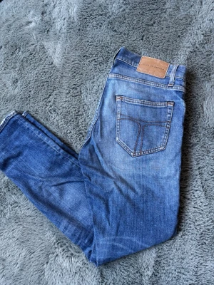 Blå jeans från Tiger of Sweden - Snygga blå jeans från Tiger of Sweden med klassisk femficksmodell och kontrastsömmar. Jeansen har en något tvättad look och är tillverkade i slitstarkt denim. Perfekta för dig som gillar stilrena och tidlösa plagg. OBS, liten fläck på ena knäet, syns knappt!