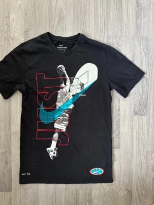 Svart Nike t-shirt med baskettryck - Snygg svart t-shirt från Nike med stort baskettryck framtill. Motivet visar en basketspelare som dunkar, med texten 'JUST' och den klassiska turkosa swoosh-loggan. Materialet är Dri-FIT som andas och känns skönt mot huden. Perfekt för dig som gillar sportig streetstyle.