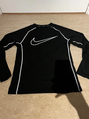Svart långärmad träningströja Nike - Svart långärmad träningströja från Nike med vit logga på bröstet och vita kontrastsömmar längs ärmar och axlar. Tillverkad i ett lätt och stretchigt material som passar perfekt för träning eller sportaktiviteter.