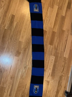Blåsvart halsduk Sirius - Snygg halsduk i blått och svart med breda ränder och Sirius klubbmärke broderat i båda ändar. Perfekt accessoar för att visa ditt stöd på matchen. Halsduken har fransar i ändarna och är tillverkad i mjukt, stickat material.