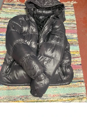 Svart pufferjacka från Brave Soul - Snygg svart pufferjacka från Brave Soul med huva och dragkedja. Jackan har två sidofickor med dragkedja och en extra ficka på ärmen. Tillverkad i glansigt material som ger en modern look. Perfekt för kalla vinterdagar.