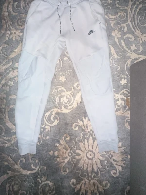Nike tech Byxor  - Snygga vita mjukisbyxor från Nike med svart logga på vänster ben och dragsko i midjan. Byxorna har ribbade muddar vid bensluten och är tillverkade i mjukt material för maximal komfort. Perfekta för en avslappnad stil.