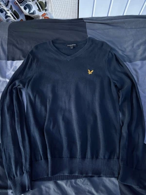 Mörkblå stickad tröja med v-ringning - Mörkblå stickad tröja från Lyle & Scott med v-ringad hals och gul broderad logga på bröstet. Tröjan har långa ärmar och ribbade muddar vid ärmslut och nederkant. Perfekt för lager på lager och stilren look. Tröjan har ett litet hål i höger armhåla bak, men kan lätt sys ihop. Pris kan diskuteras.