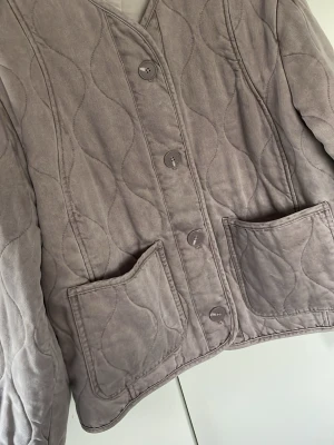 Quiltad beige grå jacka med knappar Zara  - Quiltad beige grå jacka med vågigt mönster, stora framfickor och knäppning med fyra knappar. Jackan har lång ärm och en avslappnad passform, perfekt för lager på lager. Materialet känns mjukt och ger en stilren look. Lite översized model 