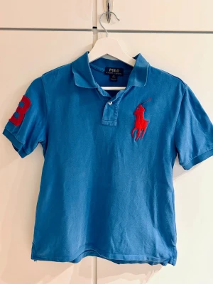 Blå pikétröja från Polo Ralph Lauren - Blå kortärmad pikétröja från Polo Ralph Lauren med röd logga på bröstet och siffran 3 på ärmen. Ungdomsstorlek 10-12/M, passar ca 10-12 år. Längd 52 cm, bredd 40 cm.