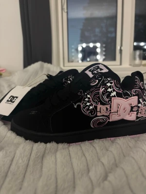 Svarta DC sneakers med rosa detaljer - Svarta sneakers från DC Shoes med stora rosa loggor och coola vita och rosa mönster på sidorna. Skorna har tjock sula, klassisk snörning och är gjorda i mockaliknande material. Perfekt för dig som gillar streetstyle och vill sticka ut med snygga detaljer.