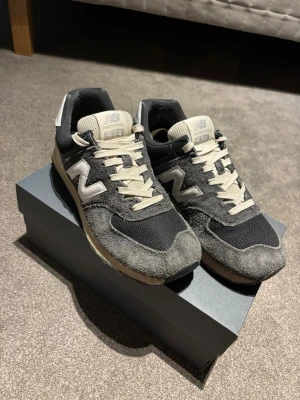 New Balance 574 mörkgrå/navy sneakers - Klassiska New Balance 574 sneakers i mörkgrått/navy med beige detaljer. Ovandelen är i mocka och mesh, med den ikoniska N-loggan på sidan och vit hälkappa. Skön retrostil och tidlös design som passar till det mesta. Använda ett fåtal gånger och är i princip helt nya! Storleken i dem är 43 men jag har vanligtvis 42-42 1/2 och dem passar mig!  PRUTBART!