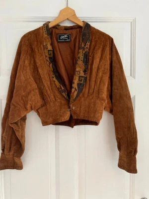 Brun kort skinnjacka med mönstrad krage - Säljer en brun kort skinnjacka från Lace med breda axlar och en unik mönstrad krage i jordnära färger. Jackan har en avslappnad passform, långa ärmar och knäppning framtill. Perfekt för dig som gillar vintage och statement-plagg.