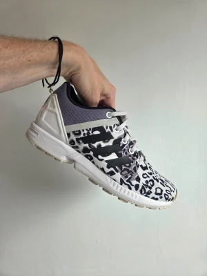 Adidas originals ZX Flux - Snygga Adidas sneakers med leopardmönster i svart, vitt och grått. Skorna har svarta ränder på sidan och vit sula. Ovandelen är i textil och syntet med perforerat grått parti vid hälen. Perfekta för dig som vill sticka ut med din stil.
