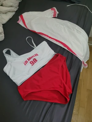 Vit och röd bikini med sportig look - Säljer en sportig bikini med vit topp och röda detaljer, texten 'Los Angeles 98' framtill. Matchande röda bikinitrosor. Perfekt för stranden eller poolen, ger en cool och ungdomlig vibe.