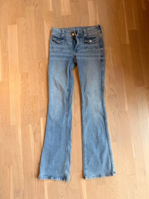 Ljusblå bootcut jeans HM - Säljer ett par ljusblå bootcut jeans med dubbla framfickor och knappdetaljer. Jeansen har en klassisk femficksdesign och utsvängda ben. Perfekta för dig som gillar retrostil och vill ha ett par bekväma jeans i mjukt denimtyg.