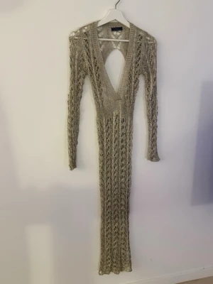 Lång beige stickad klänning - Trendig långklänning i beige stickat material med djup v-ringning och öppen rygg. Klänningen har lång ärm och ett snyggt hålmönster som ger en unik look. Perfekt för dig som vill sticka ut med en elegant och modern stil.