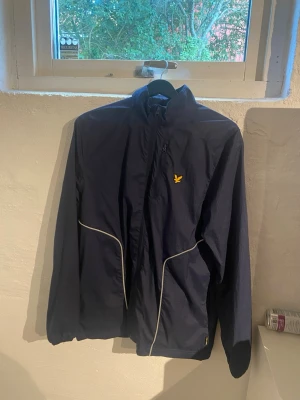 Mörkblå vindjacka från Lyle & Scott - Snygg mörkblå vindjacka från Lyle & Scott med gul logga på bröstet och vita detaljer längs sömmarna. Jackan har hög krage, dragkedja framtill och elastiska muddar. Perfekt för blåsiga dagar och sportiga outfits. Väldigt ovanlig, svår att få tag på. Därav det högre priset. Passar S och M mest S