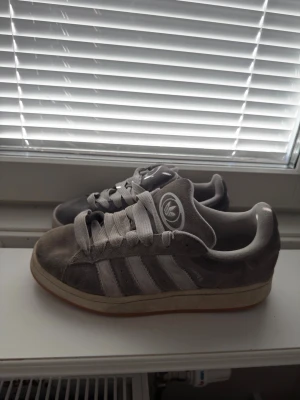 Grå Adidas sneakers i mocka - Snygga grå sneakers från Adidas med klassiska vita ränder på sidan och Adidas-logga på plösen. Skorna är låga och har en ovandel i mocka samt vita skosnören. Sulan är vit med en beige kant. Perfekta för en avslappnad streetstil.