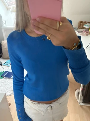 Blå topp - Snygg blå långärmad topp med dekorativa knappar vid ärmsluten. 