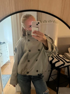 Beige kort trenchcoat med knappar - Kort beige trenchcoat med dubbelknäppning och hög krage. Jackan har vida ärmar och breda muddar