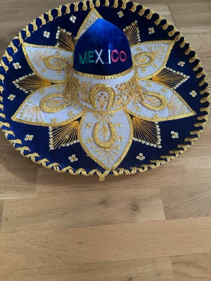 Blå mexikansk sombrero med brodyr - Säljer en blå mexikansk sombrero med detaljerad guldfärgad brodyr och vita inslag. Hatten har texten 'MEXICO' broderad framtill i flera färger. Materialet är sammet och filt, med dekorativa guldtrådar och pärlor runt hela hatten.