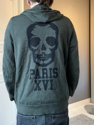 Zadig & Voltaire hoodie - Säljer en Zadig & Voltaire hoodie med stor dödskalle och texten 'PARIS XVI' på ryggen. På ena ärmen finns ett broderat motiv av en tiger och texten 'SELF CARE'. Hoodien har huva med dragsko och magficka. Perfekt för dig som gillar streetwear och coola detaljer. 