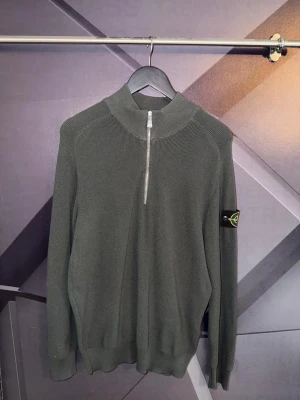 Stone island quarterzip - Snygg grå stickad tröja från Stone Island med halv dragkedja och hög krage. Tröjan har lång ärm, ribbstickade muddar och den klassiska logotypen på ärmen. Perfekt för dig som gillar stilrena och exklusiva plagg.