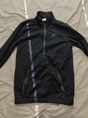 Nike half zip tröja - Svart långärmad tröja från Nike med half-zip framtill och hög krage. Tröjan har två fickor med dragkedja och diskret Nike-logga på bröstet. Perfekt för en sportig och avslappnad stil.
