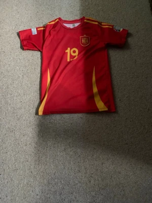 Spansk landslagströja Lamine Yamal - Röd fotbollströja med gula detaljer från Spaniens landslag, nummer 19 och namnet Lamine Yamal på ryggen. Tröjan har korta ärmar, rund hals och tryckt landslagsemblem på bröstet. Perfekt för dig som vill stötta Spanien på matchdag! Passar 12 till 13 åringar