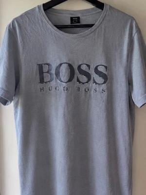 Ljusblå BOSS t-shirt med tryck - Säljer en ljusblå t-shirt från Hugo Boss med stort BOSS-tryck på bröstet. T-shirten har rund hals och korta ärmar, perfekt för dig som gillar stilrena plagg med tydlig logga. Materialet är mjukt och skönt bomull.