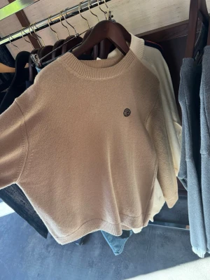 House of savoj Beige stickad tröja  - Snygg beige stickad tröja med rund hals och ribbade muddar. Tröjan har en liten broderad patch på bröstet och är tillverkad i mjukt material. Perfekt för lager på lager och enkel att matcha med olika outfits.