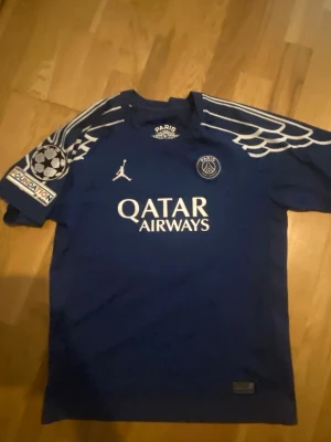 Blå PSG matchtröja med vingdetaljer - Säljer en blå Paris Saint-Germain matchtröja med vita vingdetaljer på axlarna, Champions League-märke på ärmen och Qatar Airways-tryck på bröstet. Tröjan har rund hals och är tillverkad i ett lätt funktionsmaterial som passar perfekt till fotbollsträning.