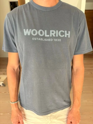 Blå t-shirt från Woolrich - Helt ny Snygg blå t-shirt från Woolrich med tryck 'WOOLRICH ESTABLISHED 1830' på bröstet. Klassisk rund hals och korta ärmar. Tillverkad i mjuk bomull som känns skön mot huden. Nypris 766 kr