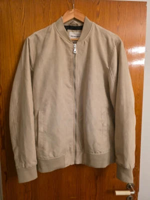 Beige bomberjacka från Jack & Jones - Stilren beige bomberjacka från Jack & Jones med dragkedja framtill och två sidofickor. Jackan har ribbade muddar vid ärmslut och midja samt en klassisk bomberkrage. Perfekt för dig som gillar en clean och enkel look.