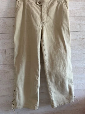 Beige Capri byxor med snörning nertill - Snygga beige Capri byxor med raka ben och unik snörningsdetalj längst ner på benen. Tillverkade i ett mjukt bomullstyg som känns bekvämt hela dagen. Perfekta för dig som vill ha en stilren men ändå lite annorlunda look.