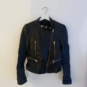 Svart bikerinspirerad skinnjacka - Snygg svart skinnjacka med asymmetrisk dragkedja och quiltade detaljer på ärmarna. Jackan har tre framfickor med dragkedja och hög krage med tryckknapp. Perfekt för en cool och trendig look.